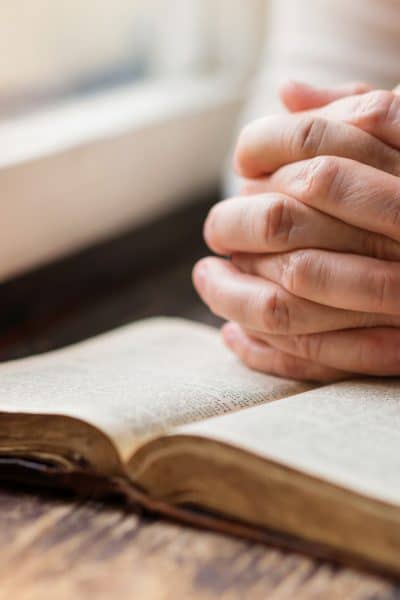 Woman by window with hands praying over a Bible